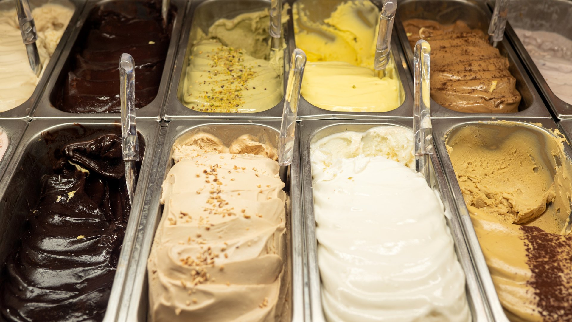 Variety of colorful ice cream flavors in metal containers at a dessert shop.