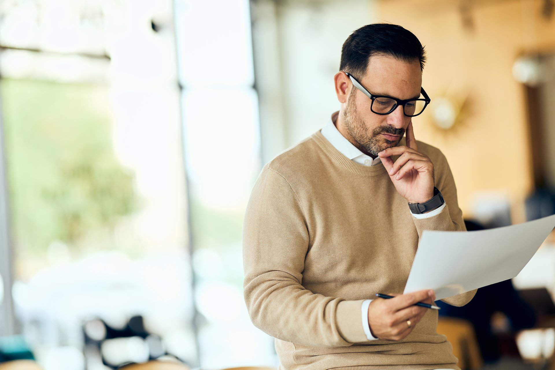 Professional man in a beige sweater reads a document in a bright office, focused and thoughtful.