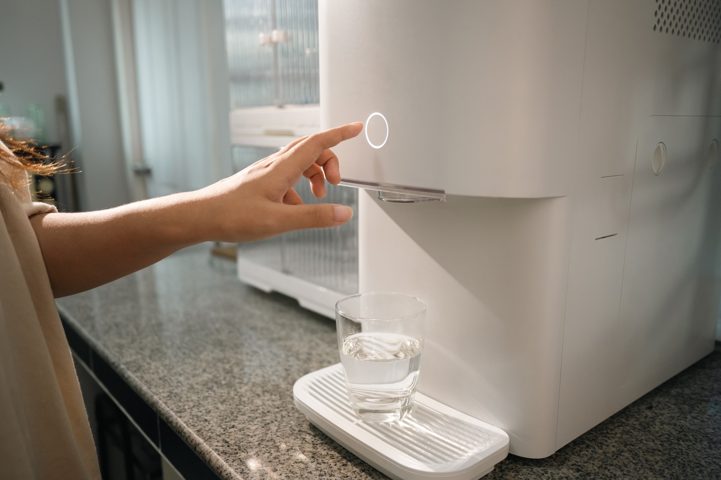 Young woman pressing button on a modern water purifier, filling a glass with fresh water.
