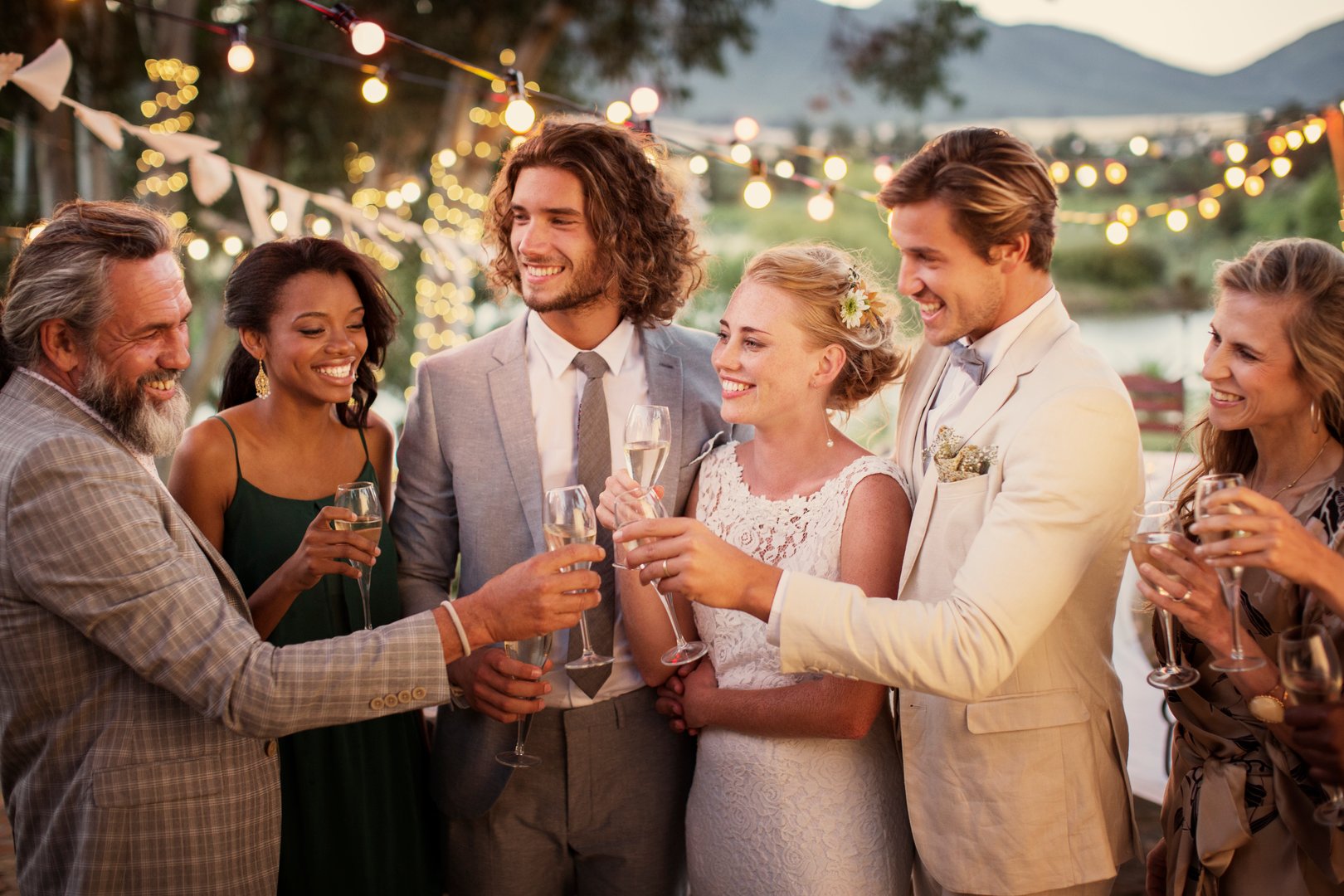 Group of people celebrating, raising glasses at an outdoor event with string lights and mountains in the background.