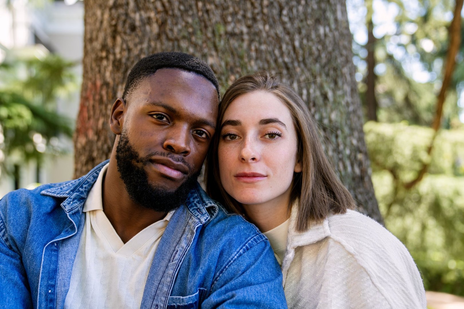 Young multiethnic couple leaning against a tree in a park, looking at the camera with calm expressions, exuding love and connection