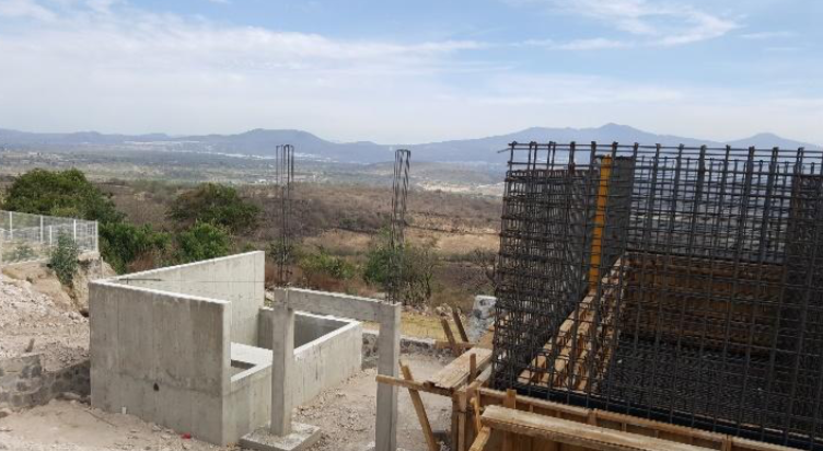 Construction site with concrete structures and steel reinforcements. Mountains and trees visible in the background.