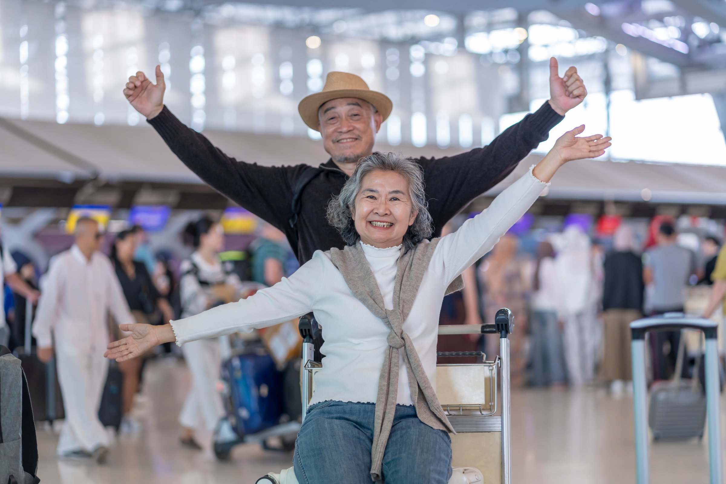 happy asian senior woman sitting on luggage that put on trolley,raising hands up,her husband standing behind in airport,excited and ready to travel