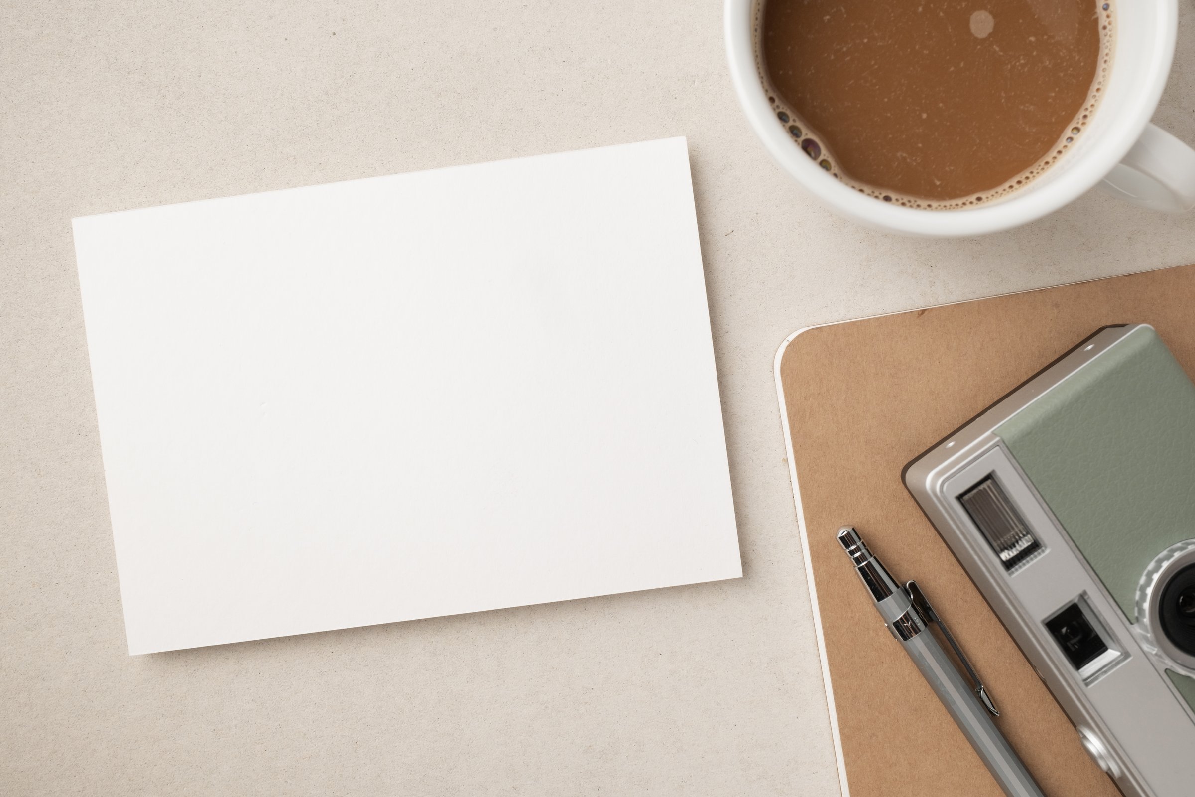 Blank white greeting card or invitation with coffee cup, camera, notebook and pencil on beige background. Mockup presentation. Flat lay. Top view with copy space. Modern minimalist style.