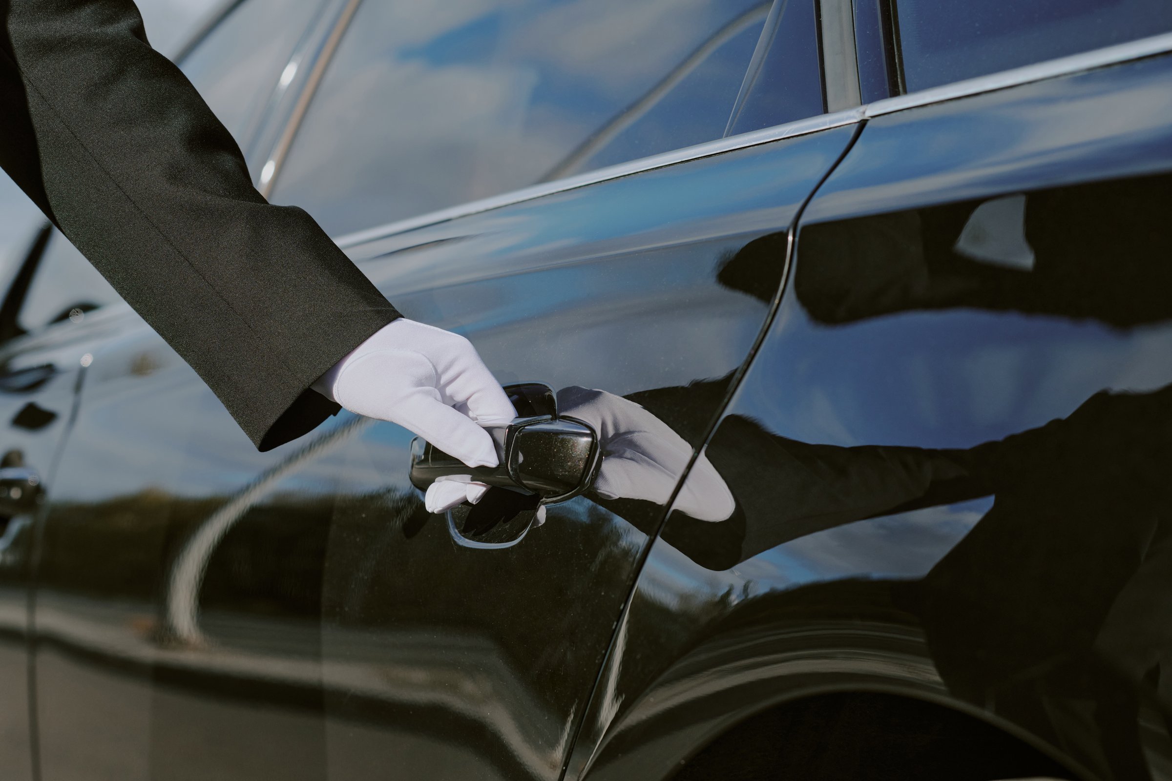 Caucasian middle aged man wearing glove opening car door, only hand and part of arm visible, luxury vehicle reflecting surroundings, professional chauffeur service concept