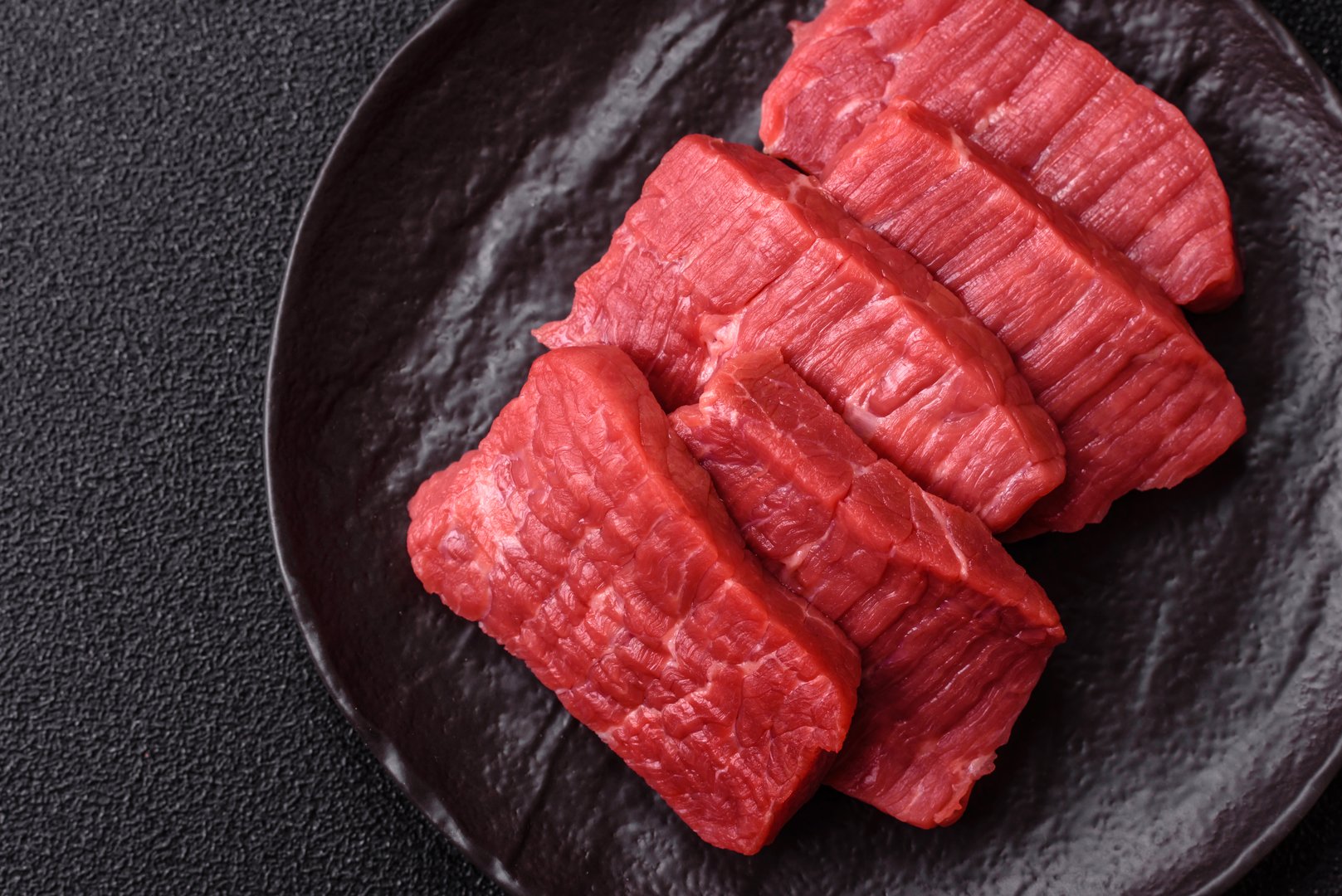 Raw beef tenderloin cut into slices with salt and spices on a dark concrete background
