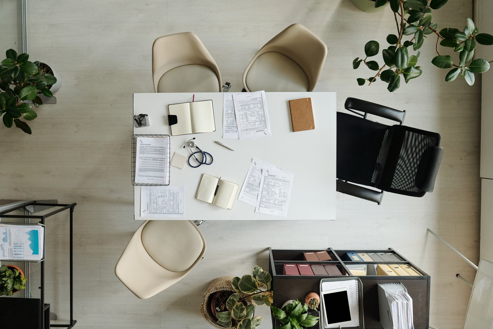 Top view showing empty office meeting table with scattered documents, open notebooks, eyeglasses, pen, and surrounding chairs, indoor workspace with potted plants and organized shelves