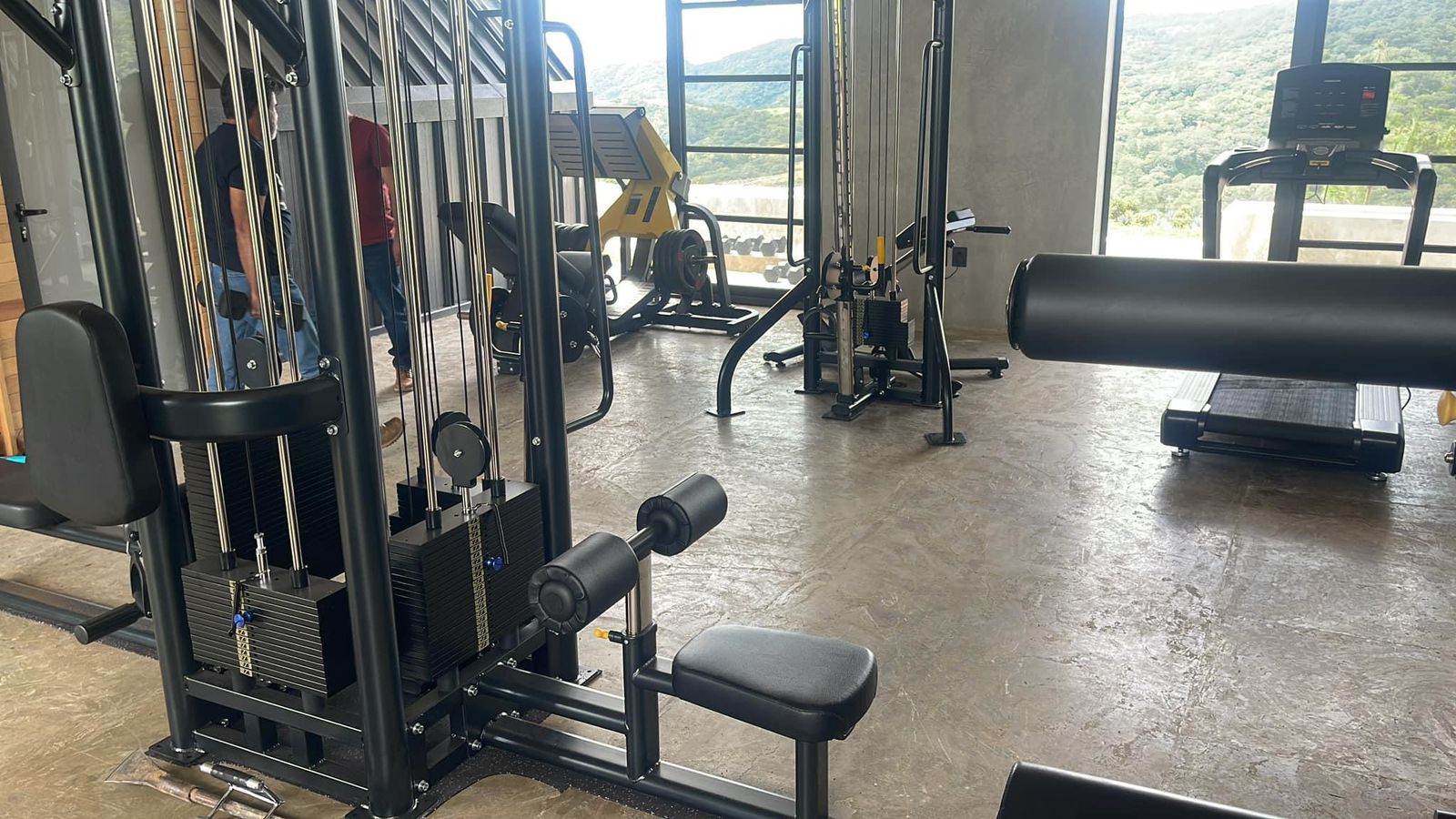 Modern gym with exercise equipment including a leg press machine, treadmill, and cable machine. Large windows with scenic view.