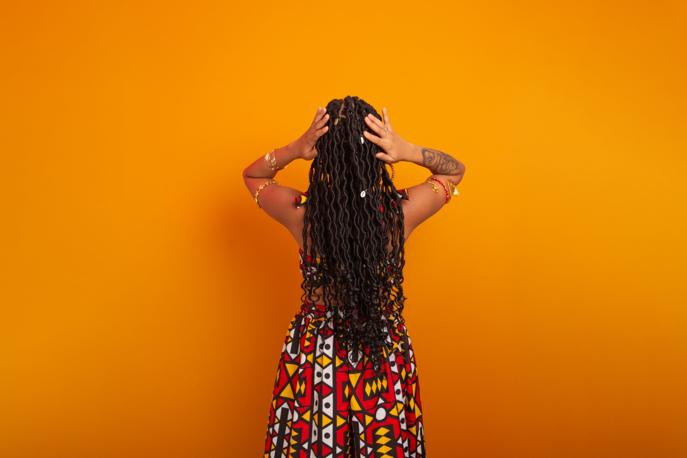 Beautiful young woman with braids in her hair, standing with her back turned, wearing traditional clothing, posing for the camera. Isolated on a mustard-colored background. Confident and calm person.