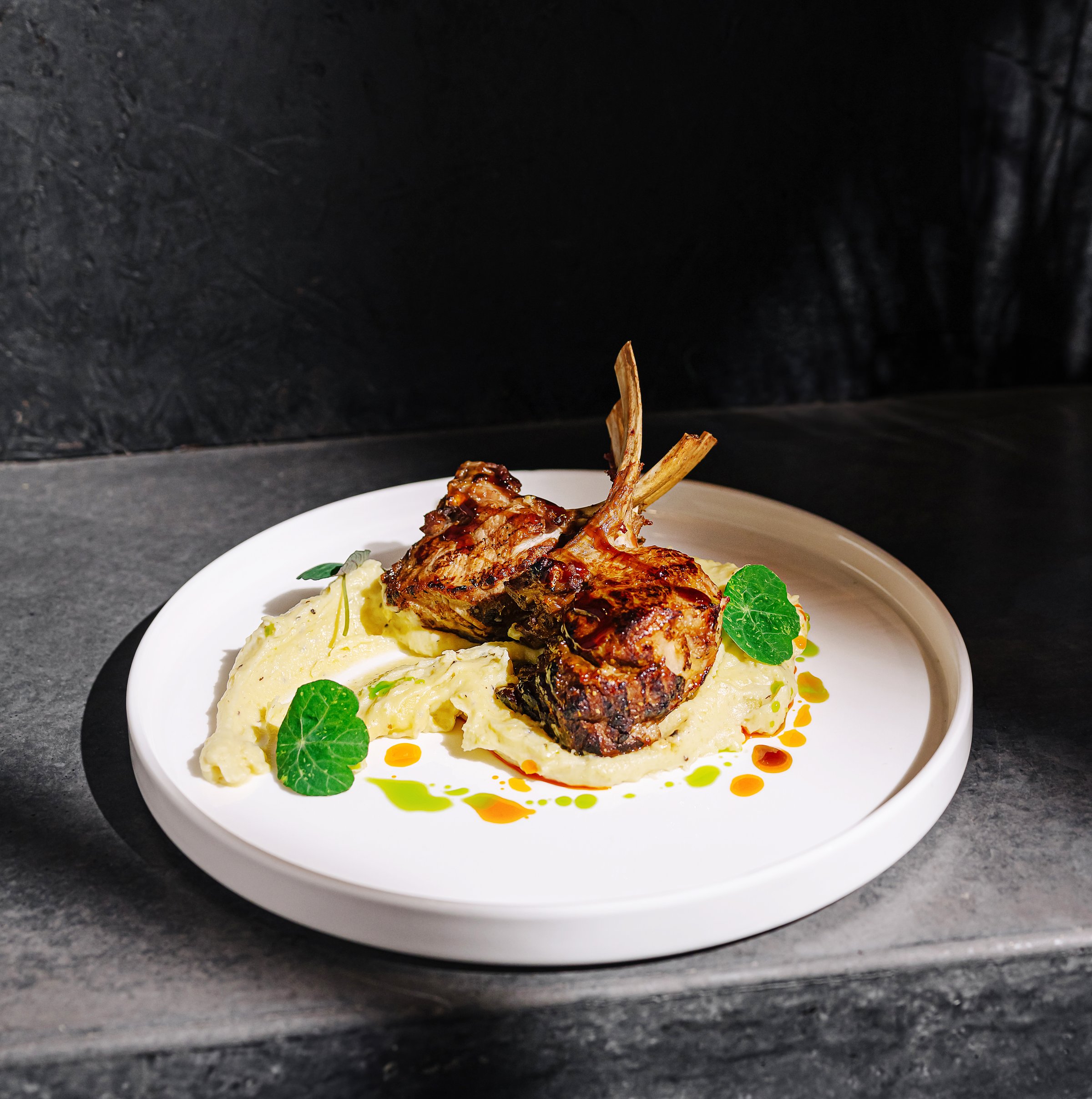 Delicious grilled lamb chops are plated beautifully over a rich creamy puree, garnished with fresh leaves and a drizzle of sauce, showcasing culinary artistry in a cozy restaurant.