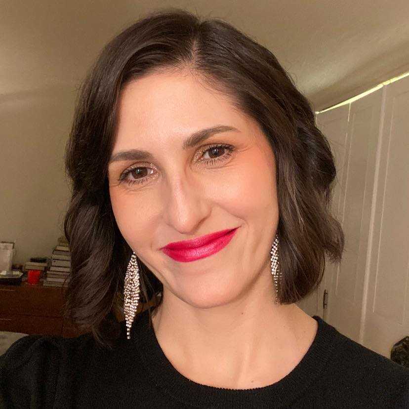 Person with shoulder-length dark hair, wearing red lipstick and dangling earrings, smiles in a warmly lit room.