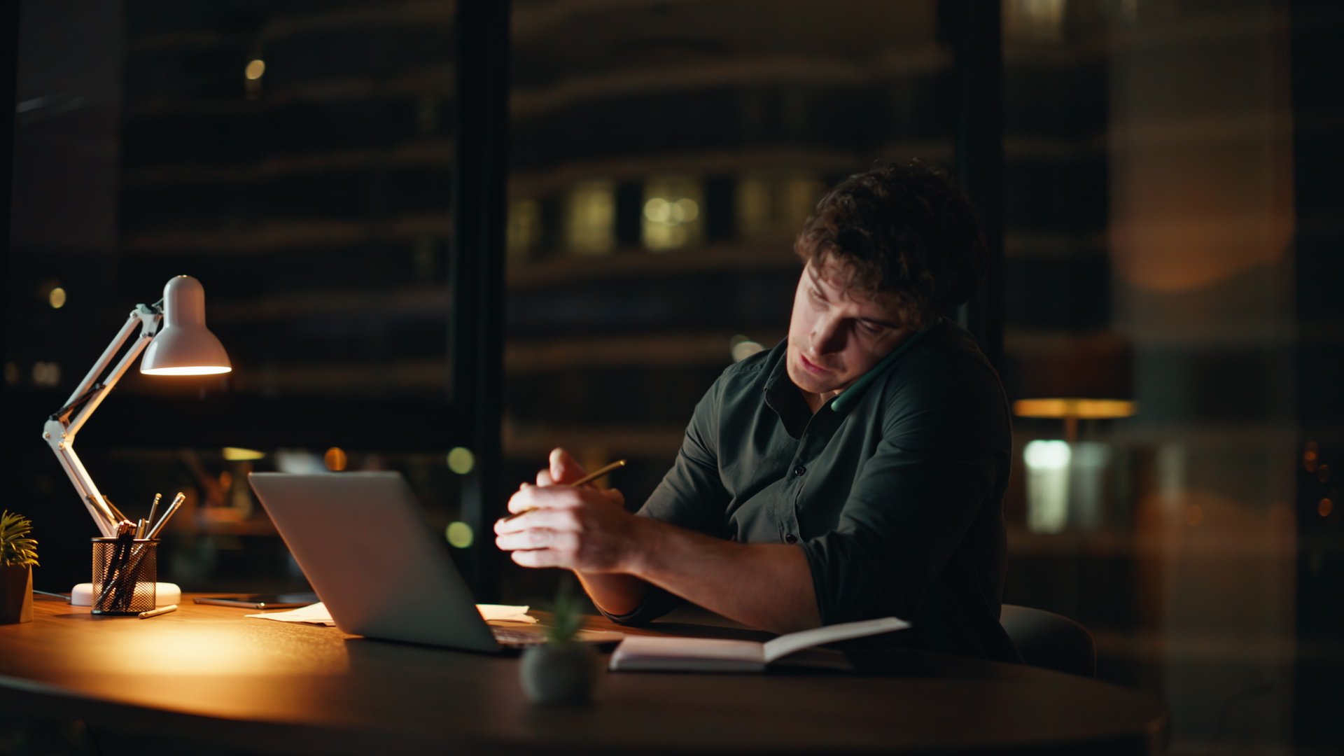 Overloaded freelancer calling cellphone looking laptop at night office. Busy businessman talking smartphone working computer late evening. Multitasking manager discussing business task in conversation