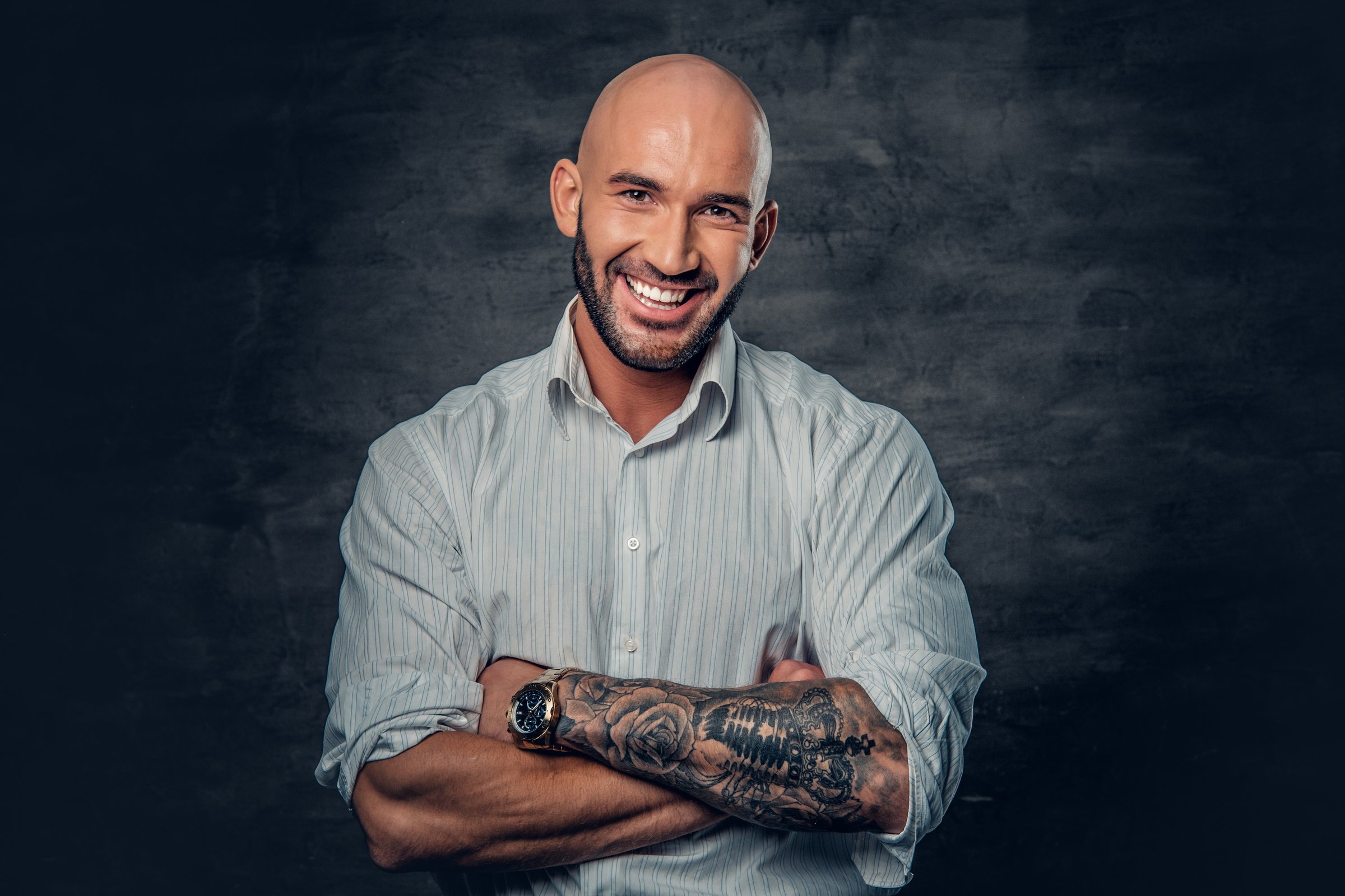 Portrait of shaved head male in a white shirt with tattooed crossed arms.