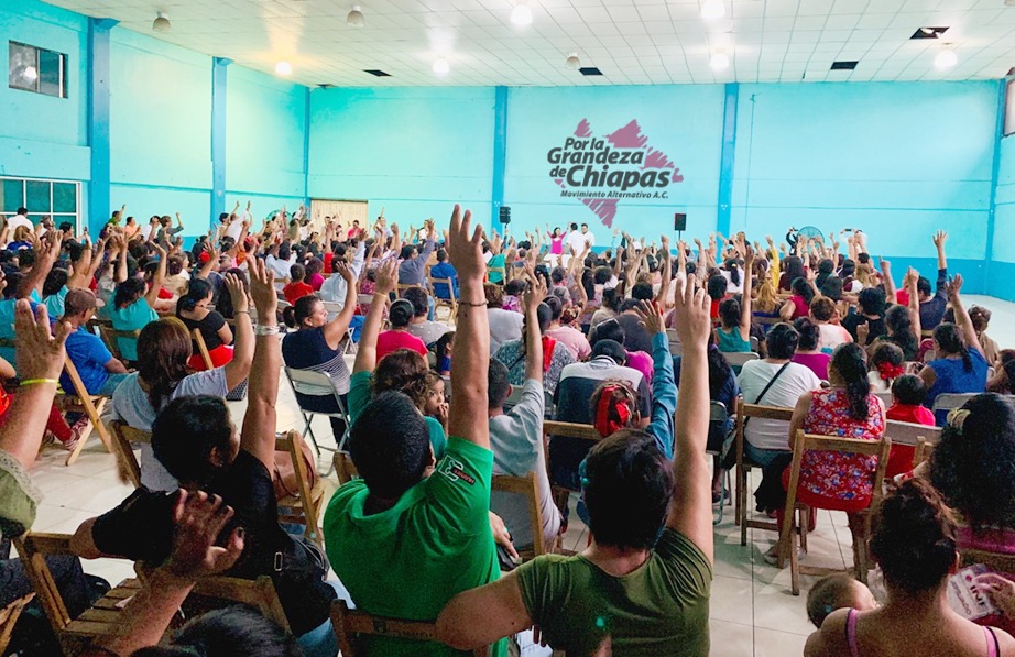 A large group of people seated in a hall raising their hands in a meeting, with a sign on the wall reading Por la Grandeza de Chiapas.
