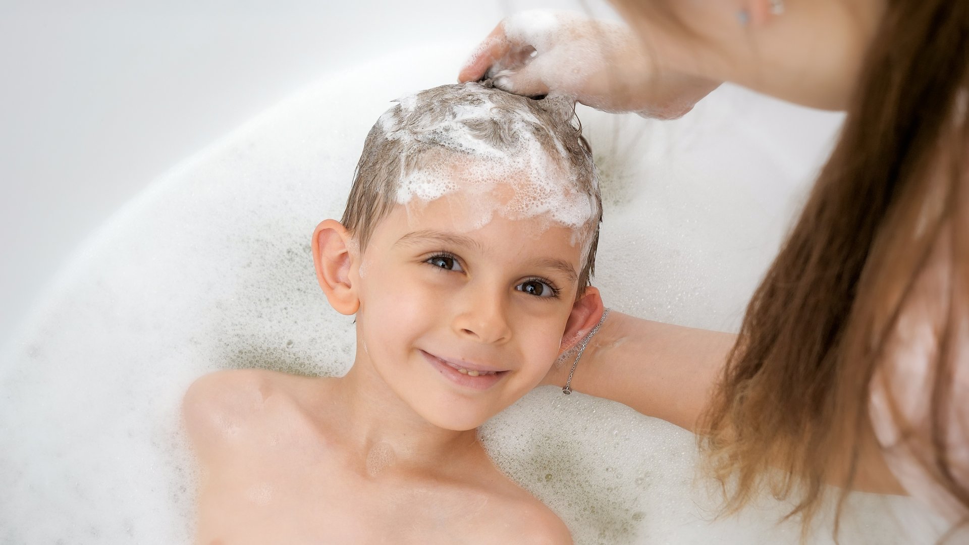 Young mother applying shampoo and washing head of her smiling son in bath.
