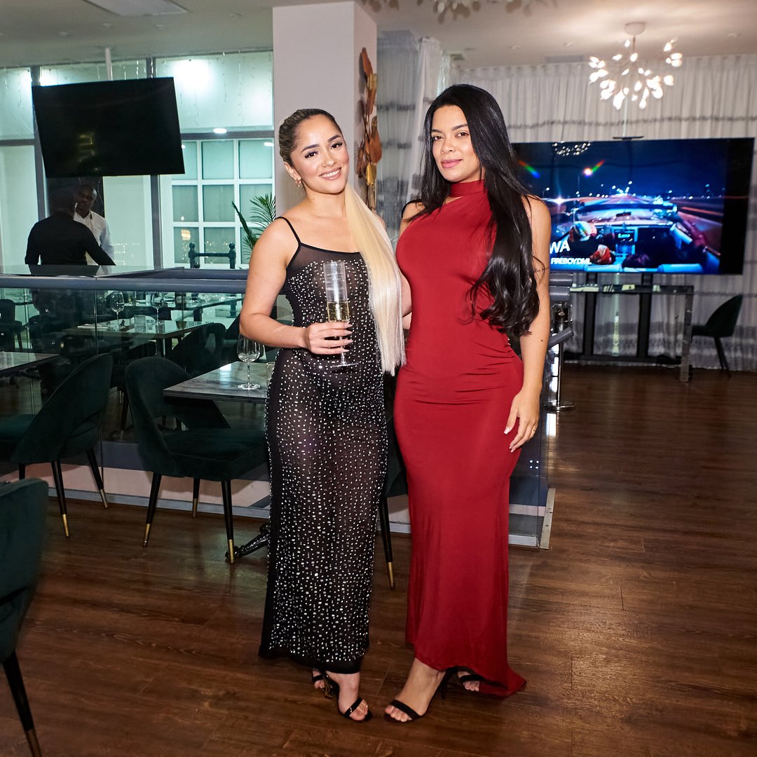 Two women pose in evening gowns; one in black sparkling dress, the other in red. They're in a modern indoor setting.