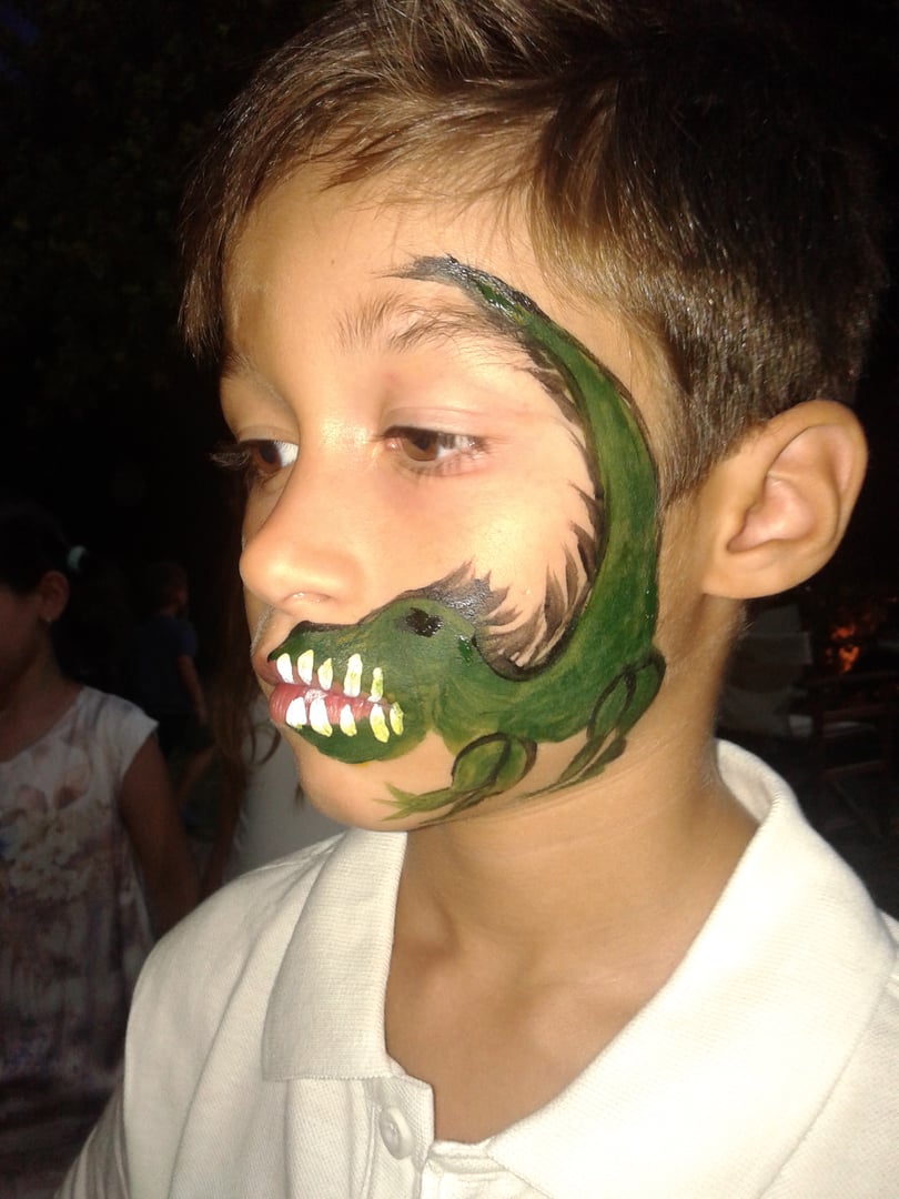 Child with a dinosaur face paint design on the cheek, wearing a white shirt, looking to the side.