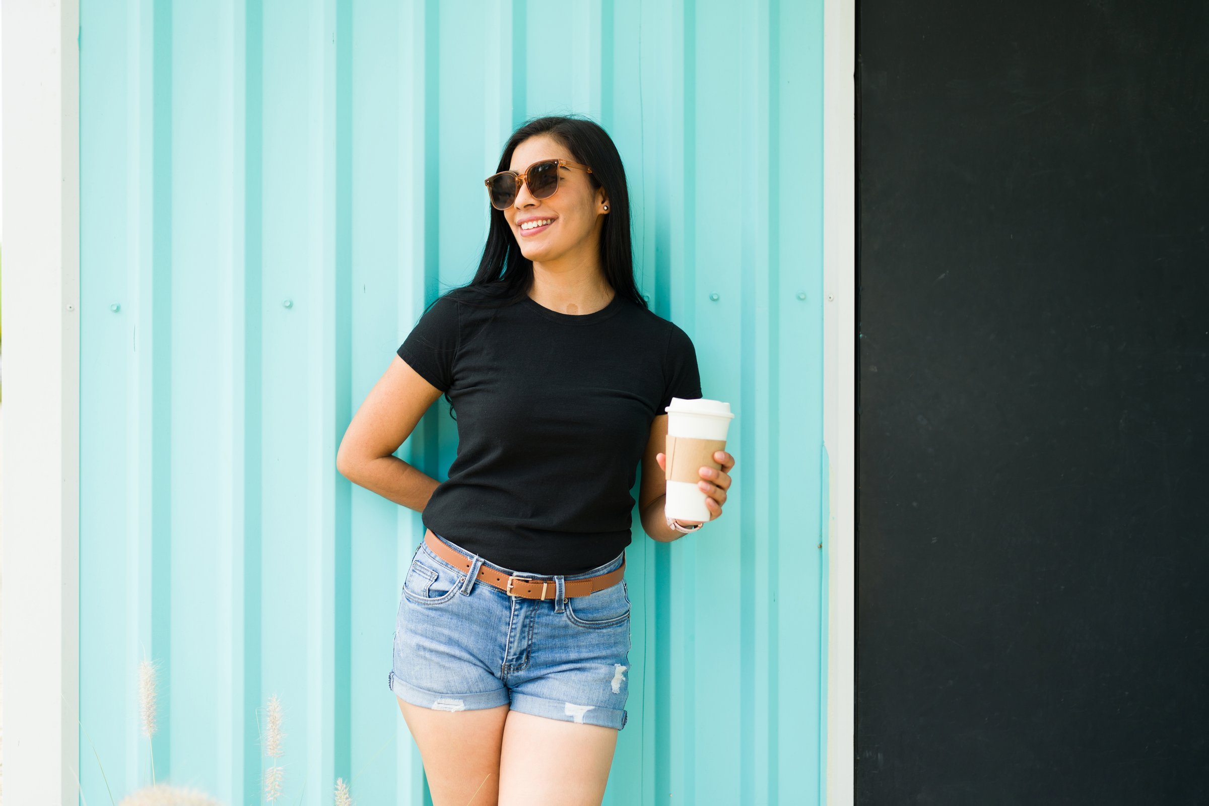 Smiling Hispanic woman in black t-shirt holding coffee cup stands confidently against a teal background with sunglasses on a sunny day