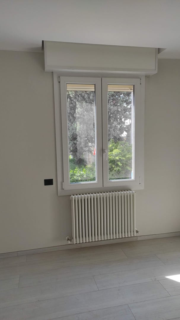 A white window with a radiator beneath it, set in a light-colored wall, overlooking a garden view.
