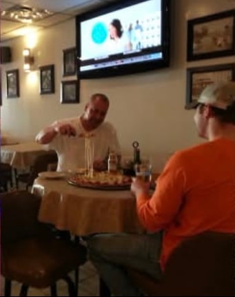 Two men dining at a pizzeria table, one pulling a slice of pizza with cheese stretching, television on the wall in the background.