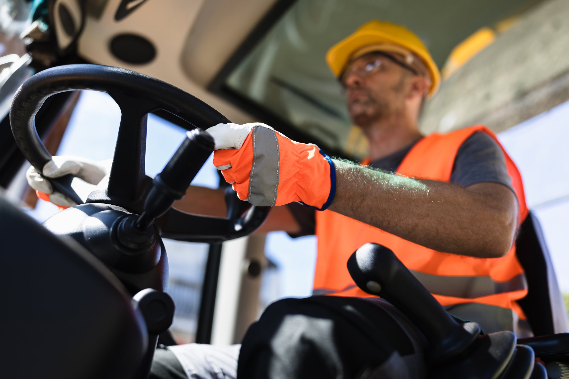 A construction worker in a reflective vest and safety gloves is focused on operating a heavy machine at a bustling construction site. The sun illuminates the area.