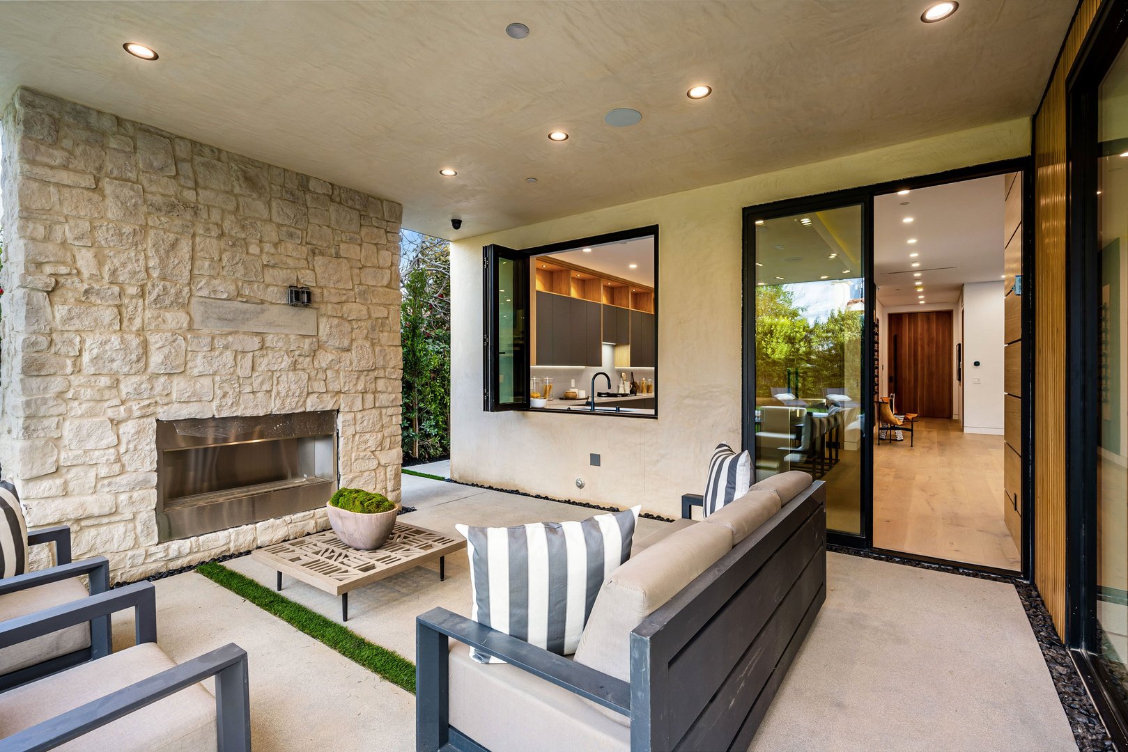 Modern outdoor patio with stone fireplace, stylish furniture, and view into contemporary kitchen and living area.