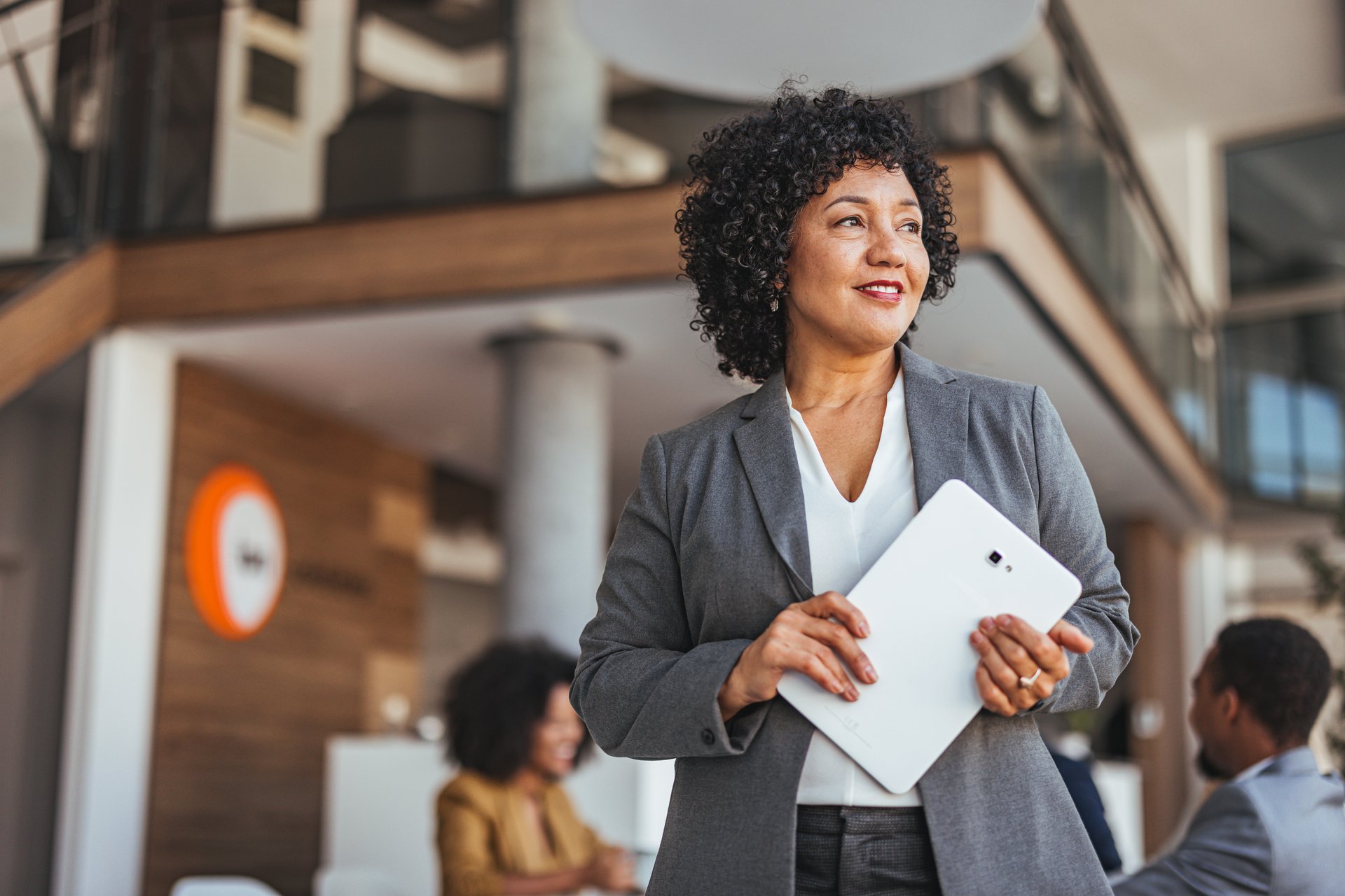 Professional woman in a formal suit holding a tablet in a modern office, exuding confidence and leadership, with a focus on teamwork and corporate success.