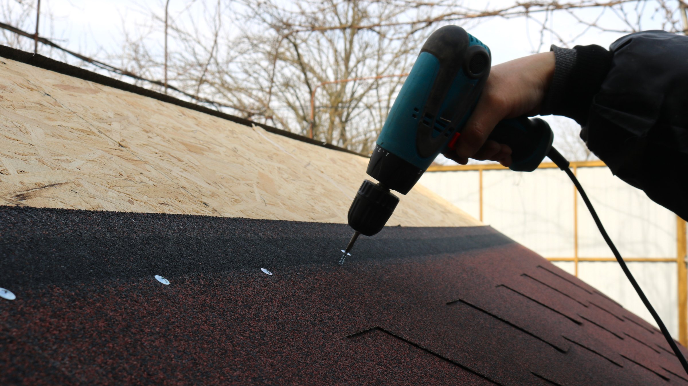 a hand in warm work clothes with a blue electric screwdriver screws fasteners into the roof structure of a low building, connecting soft tiles and an OSB sheet