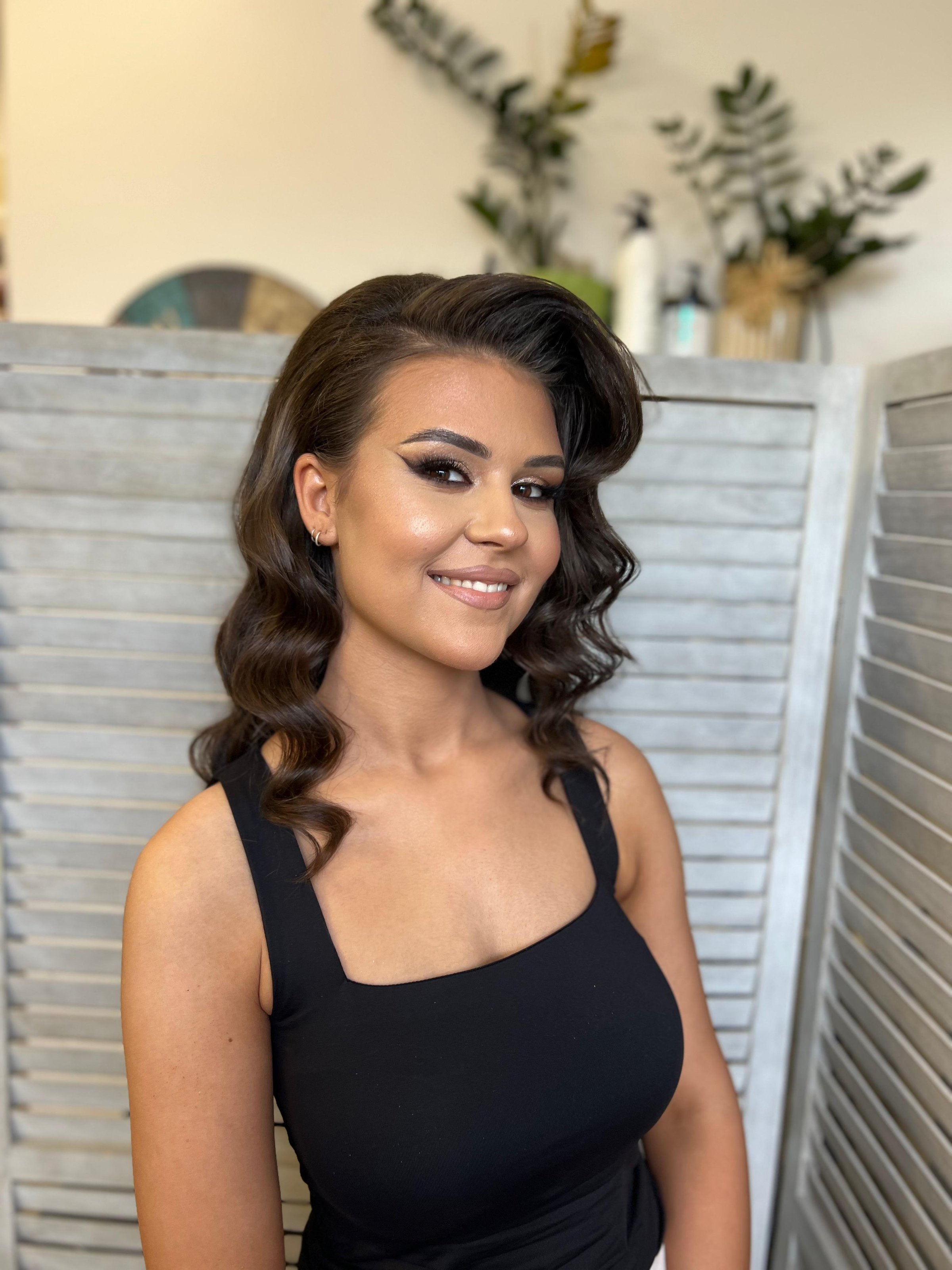 Woman with wavy hair in a black dress smiling, standing indoors with a wooden folding screen and plants in the background.