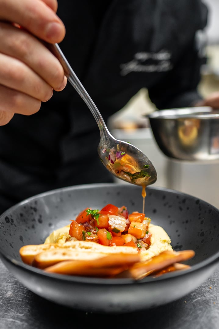 A chef skillfully plating a dish, adding a vibrant tomato topping to a smooth hummus base, showcasing culinary artistry in an elegant black bowl.