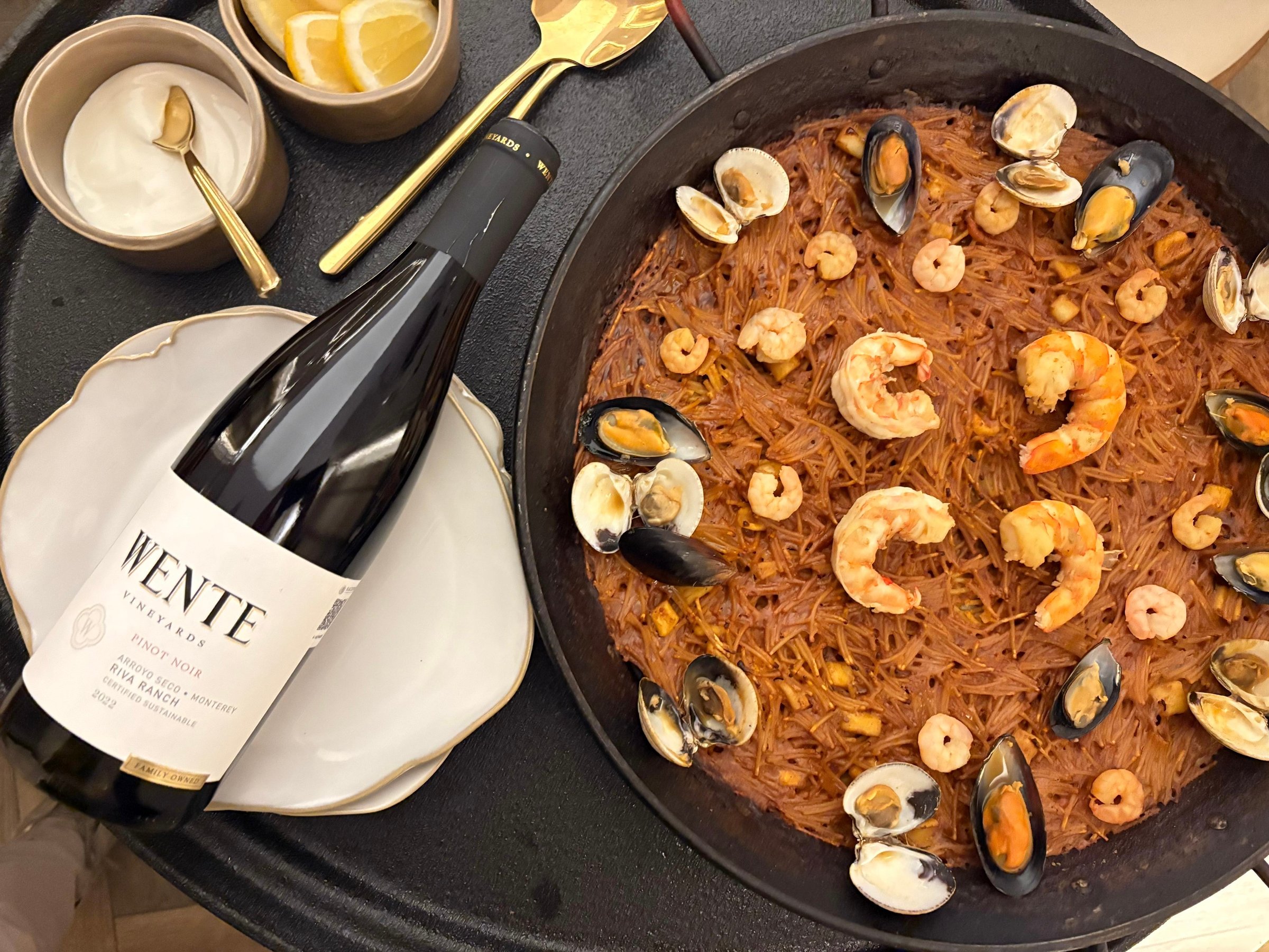 Seafood paella in a pan with shrimp, mussels, and clams, accompanied by a bottle of white wine on a table.