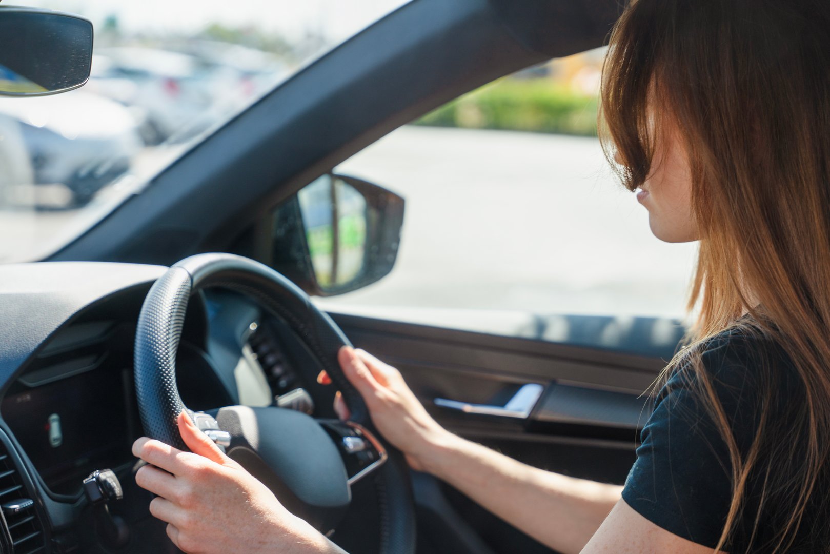 A woman is driving her vehicle in a bright parking lot, holding the steering wheel with a focused expression while sunlight shines through the car window.