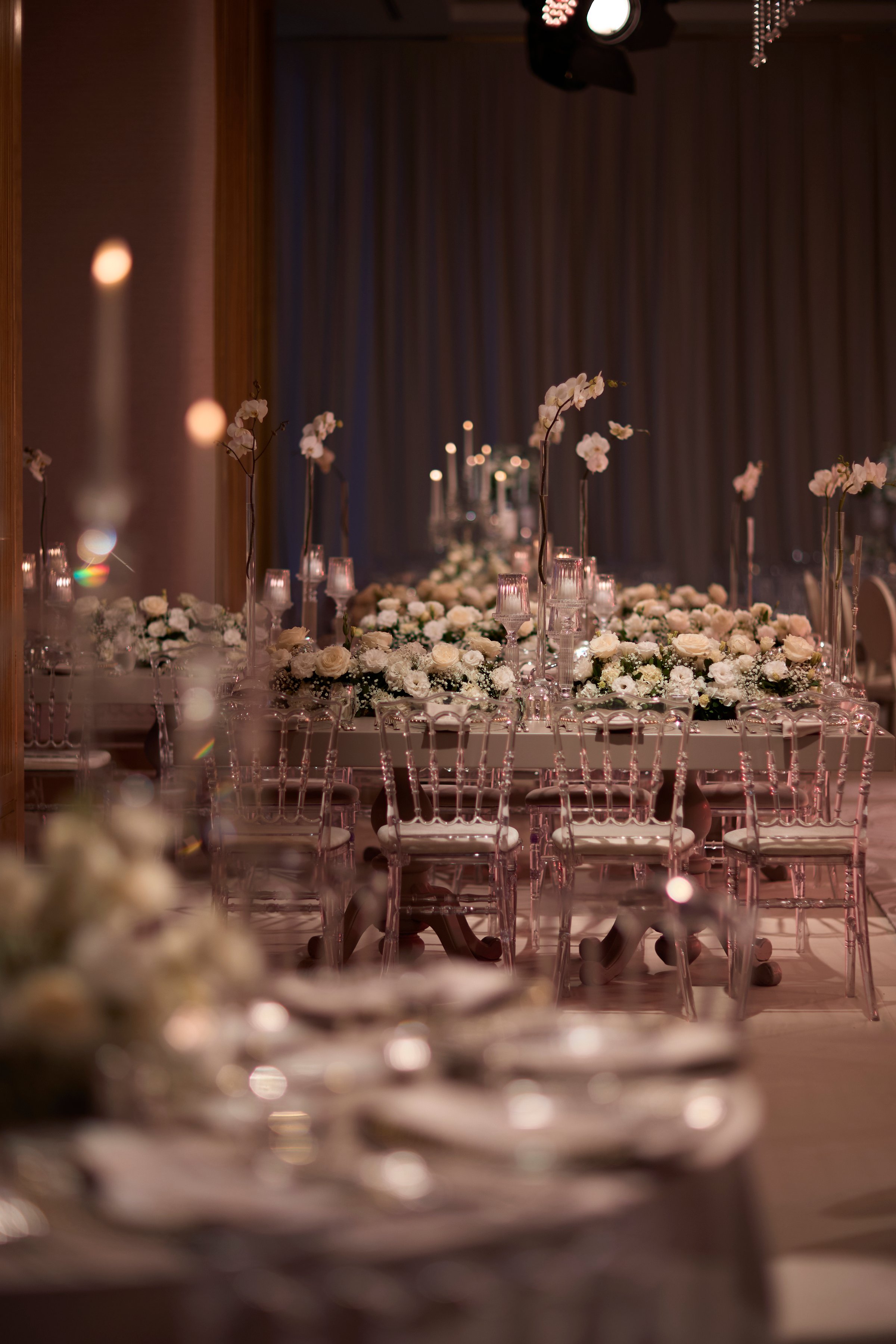 A beautifully arranged wedding reception table adorned with delicate white and cream flowers, transparent chairs, and soft candlelight. This sophisticated setting captures the essence of luxury and romance, perfect for wedding, event planning, or lifestyle projects.
