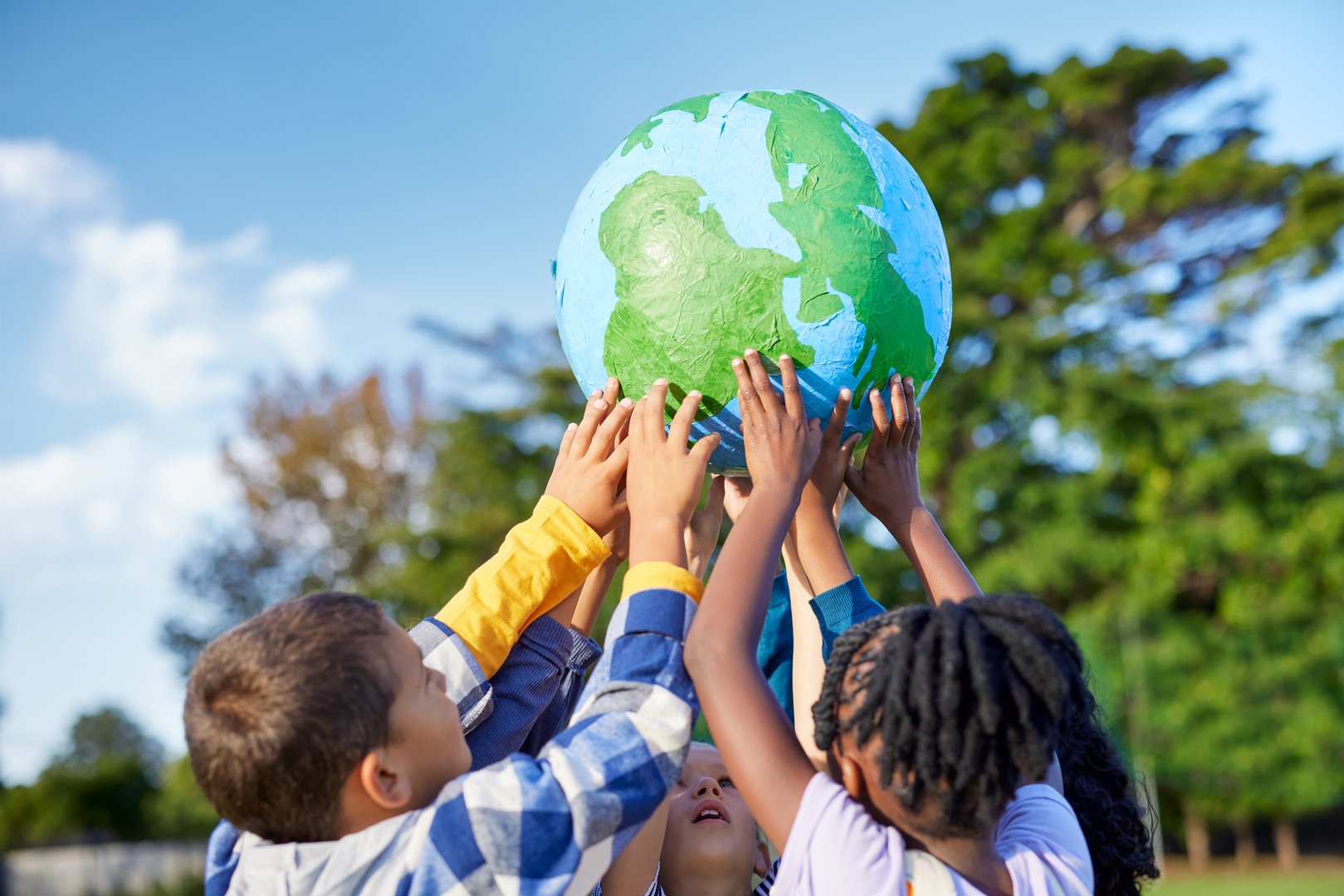 Group of multiethnic children holding model of earth in park with copy space. Primary students holding a big handmade earth model and raising it high. Hands of school children holding green world for save the planet, earth day, global peace and unity concept.