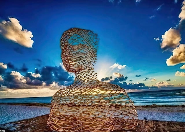 A wire sculpture of a human form stands on a beach at sunset, with clouds and the ocean in the background.