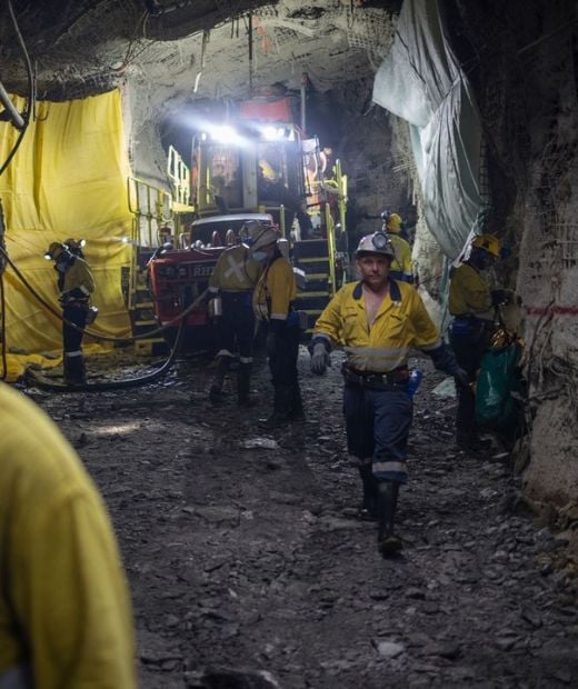 Miners in yellow uniforms work inside a dimly lit underground mine with machinery and yellow safety barriers visible.