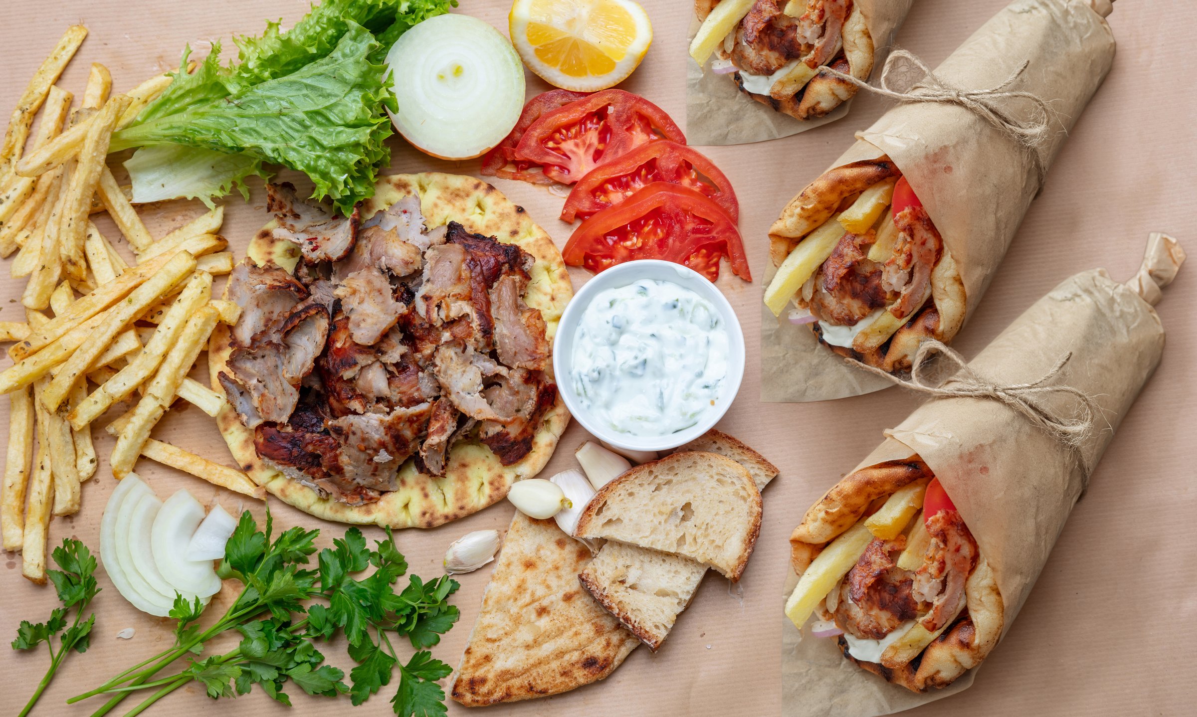 Greek gyros wraps and fresh ingredients arranged on a rustic table under natural daylight, showing pita bread, grilled meat, fries, vegetables, and tzatziki sauce in a warm inviting setting.