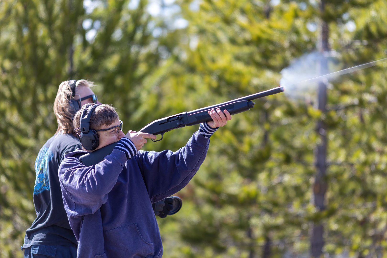 Side view of two young men shooting a 12 gauge shotgun.  Shooting clay targets. Bullet trace.  Gun powder gases coming out of barrel.