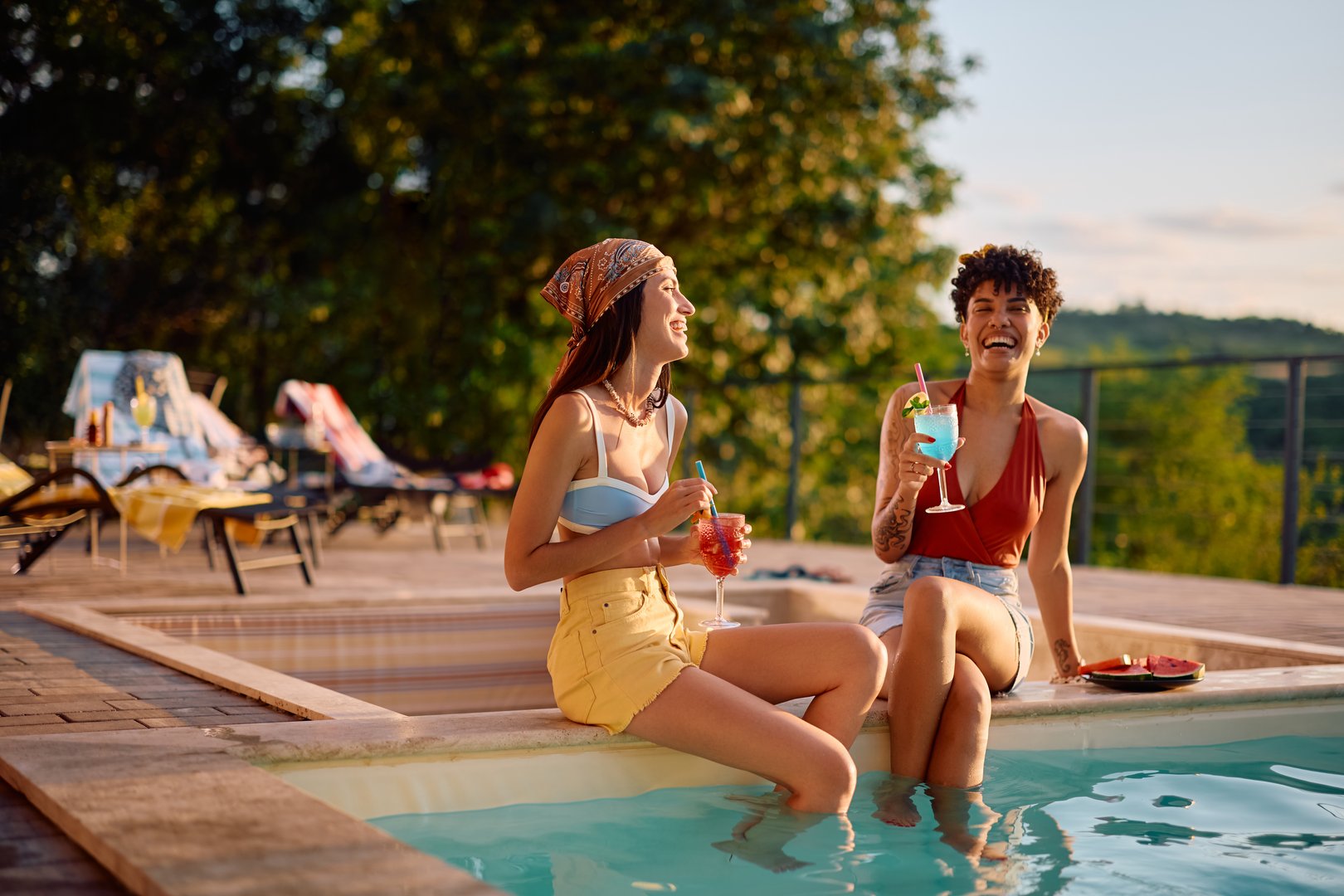 Happy female friends drinking cocktails and laughing while enjoying in summer day at swimming pool. Copy space.