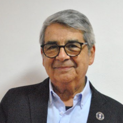 Elderly person with glasses, gray hair, wearing a suit and light blue shirt, smiling against a plain background.