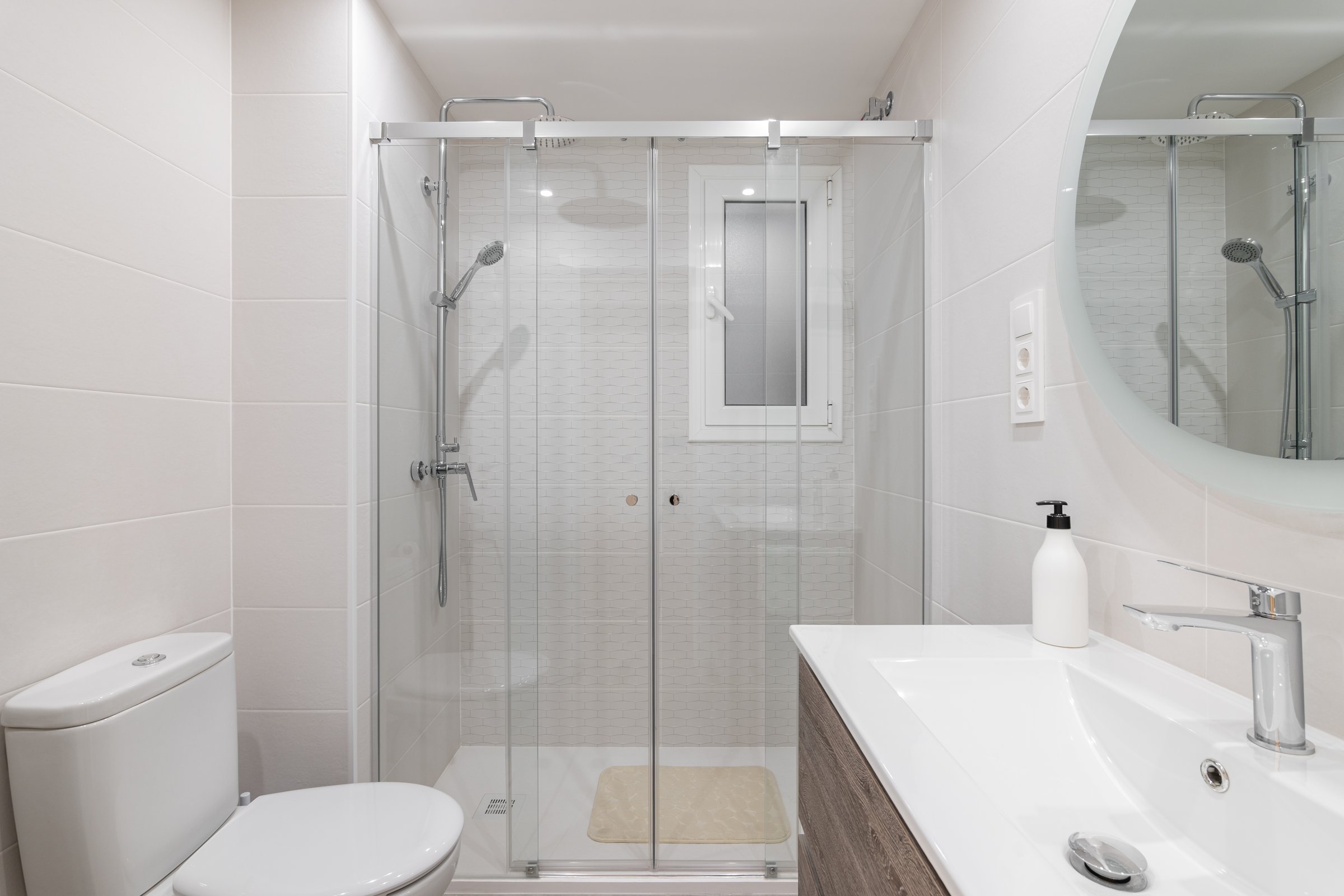 Spacious white ceramic tile bathroom is illuminated by bright white natural light. Large round mirror on the wall reflects the shower area with glass railing and metal fittings