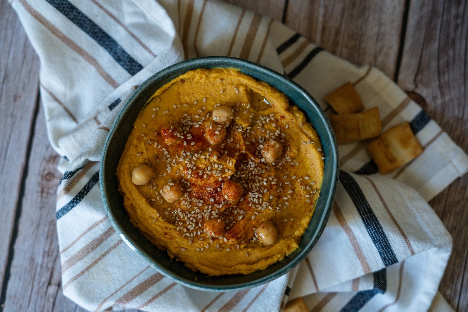 A creamy and vibrant pumpkin hummus garnished with chickpeas, presented on a dark background that accentuates the warm, rich colors of the dish. The rustic presentation is enhanced by a simple cloth, adding a cozy and earthy feel to the image.
