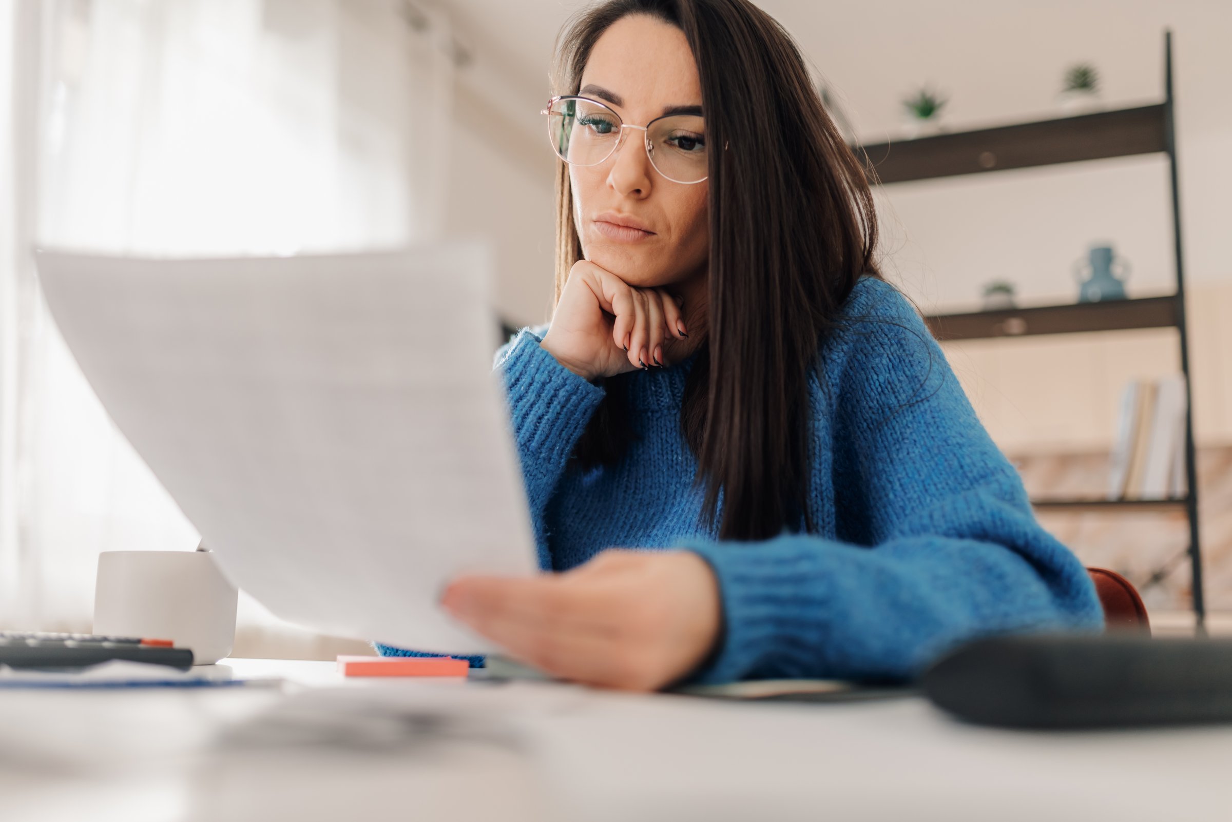 Young woman wearing glasses reviewing paperwork, managing home finances, calculating expenses, and planning budget
