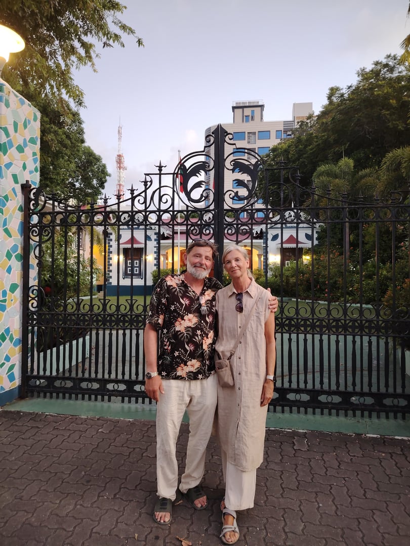 A smiling couple poses in front of a decorative iron gate with a building and trees in the background during daylight.