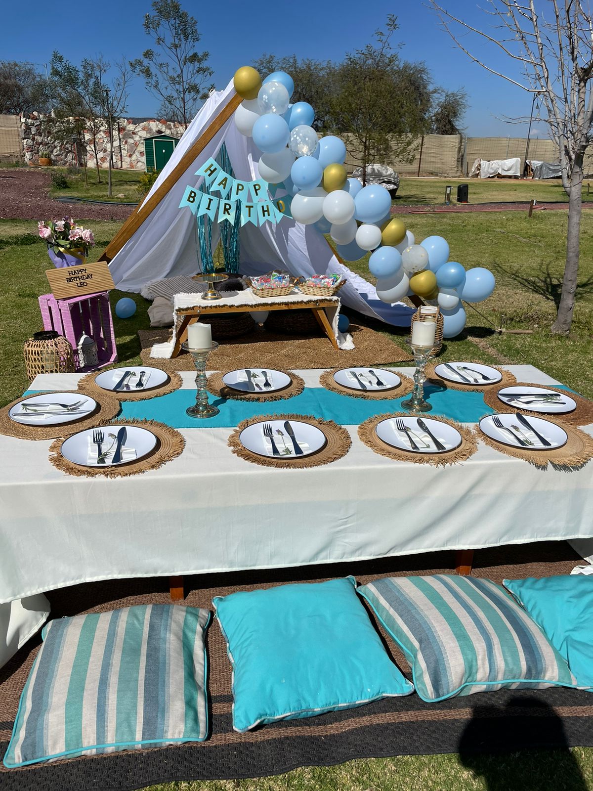 Outdoor birthday setup with a tent, balloons, and a picnic table with place settings and teal cushions.