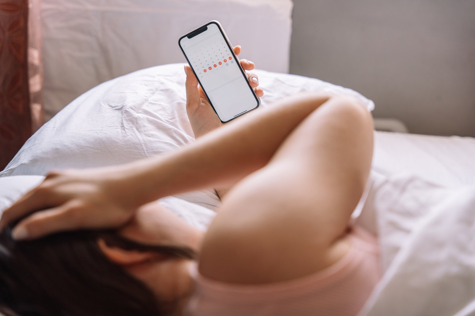 The girl has stomach pain and lies in bed holding a phone with a calendar in her hand.