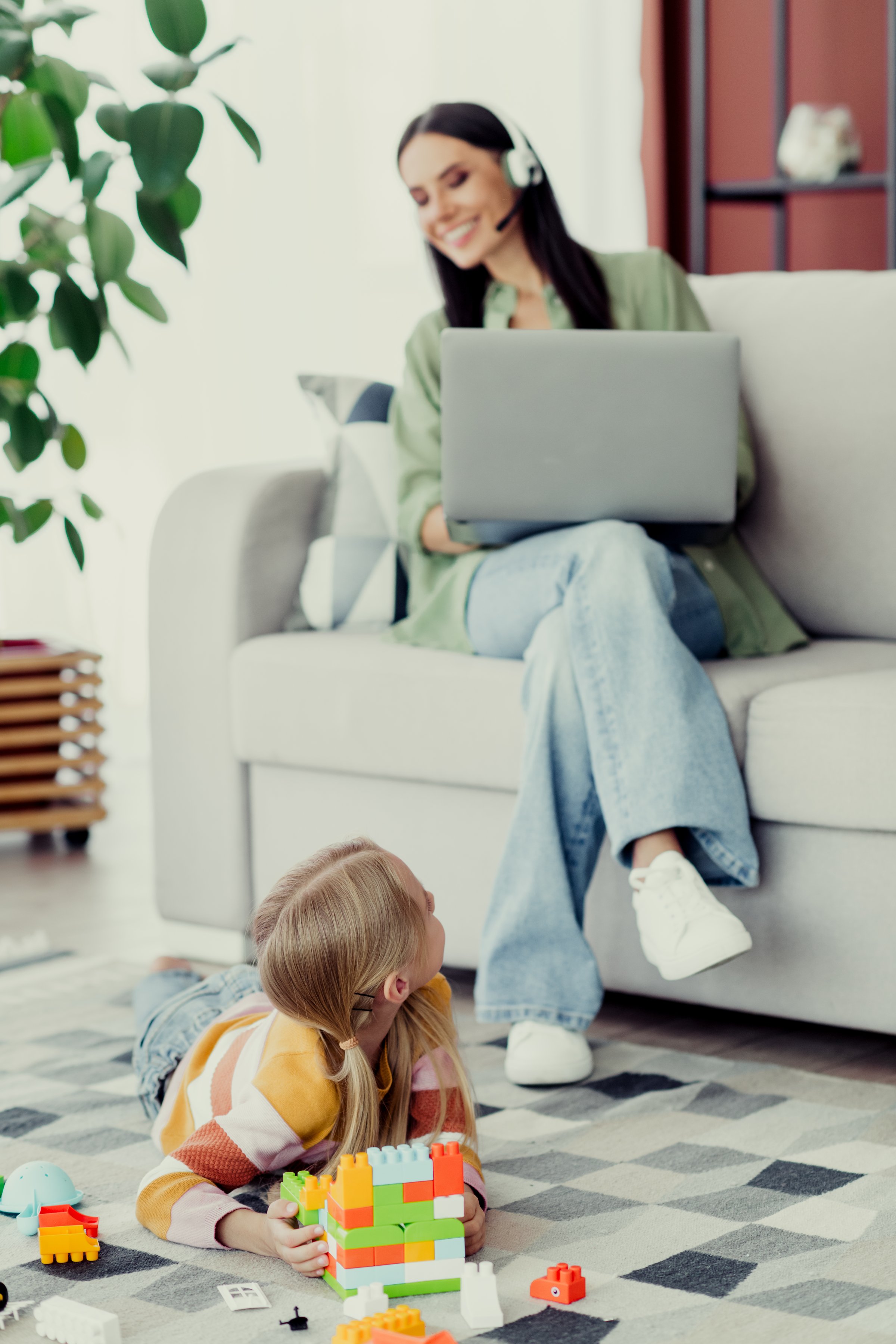 A cheerful mother works remotely while her young daughter builds with colorful blocks, creating a warm scene in a homely living room atmosphere.