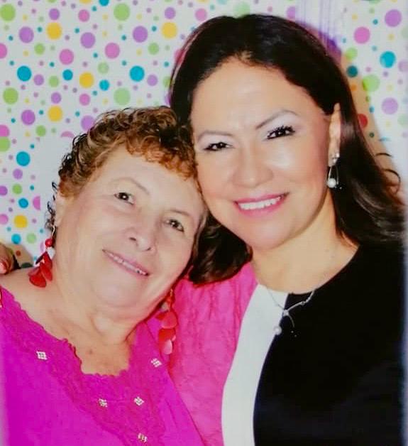 Two women smiling, one older with curly hair, the other younger with dark hair, posing in front of a colorful polka dot backdrop.