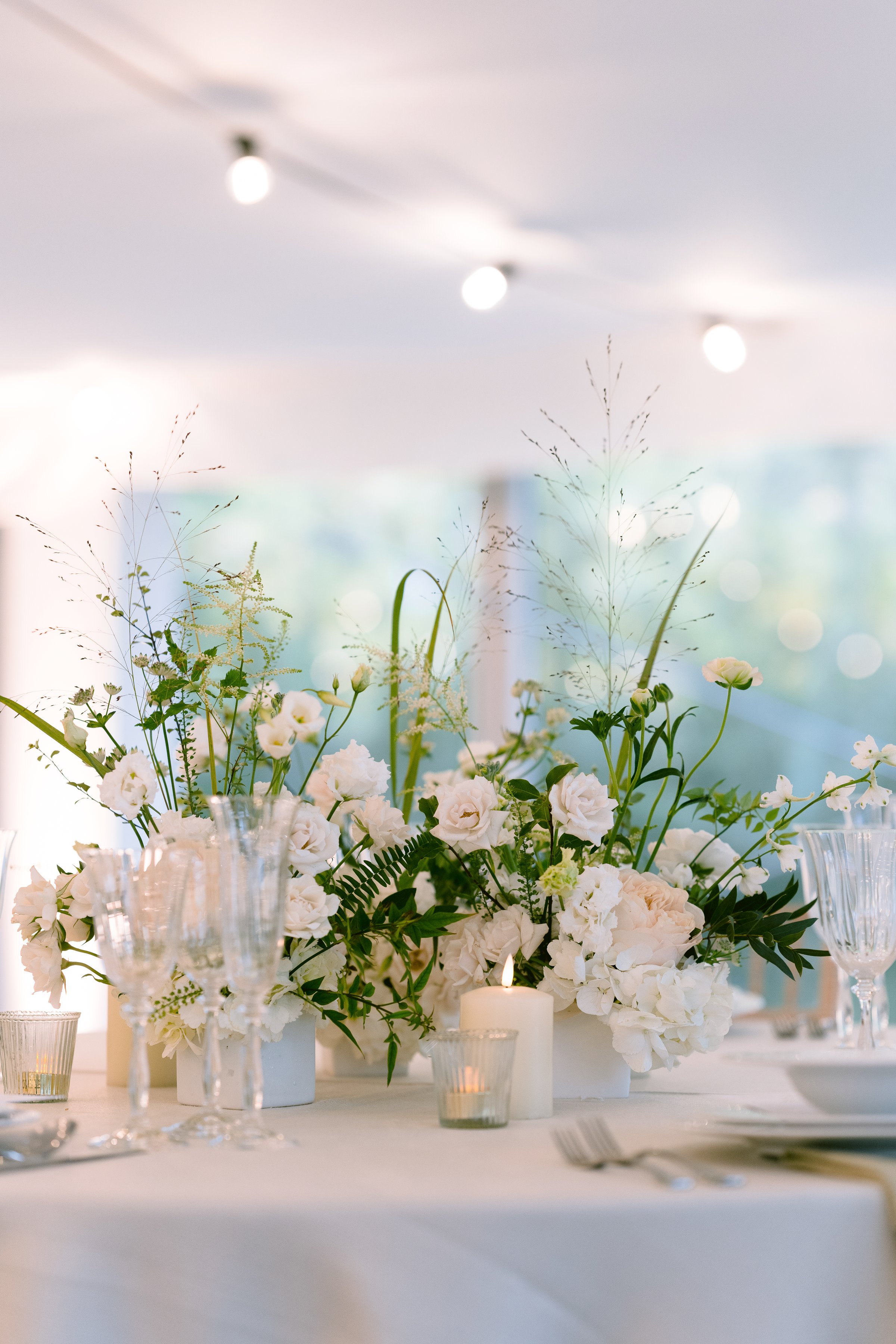 Elegant floral decoration on the back of a light upholstered chair at an indoor wedding ceremony. The arrangement features soft blush and ivory roses, subtle greenery, and a trailing silk ribbon, creating a romantic and sophisticated detail. Set within a refined indoor venue with warm lighting and a clean, minimalist aesthetic, the scene exudes timeless charm—perfect for modern, classic, or intimate wedding inspiration