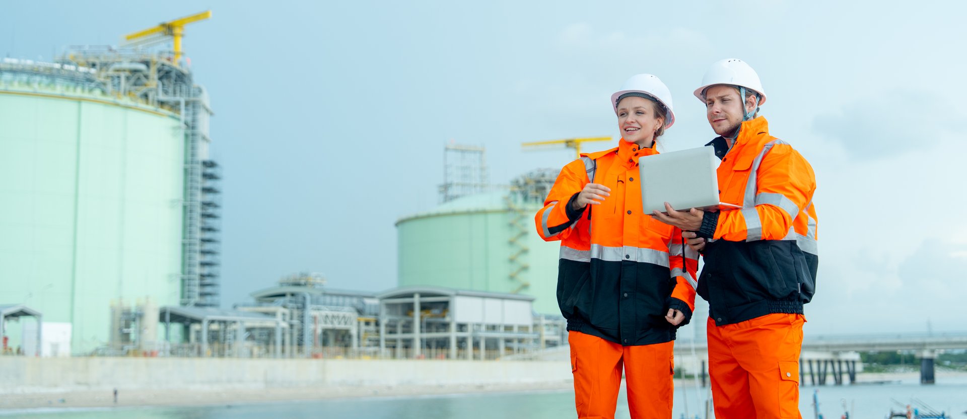 Petrochemical or gas factory workers or technician work in front of tank in factory with one woman hold walkie talkie and one man hold laptop and look to woman pointing direction.